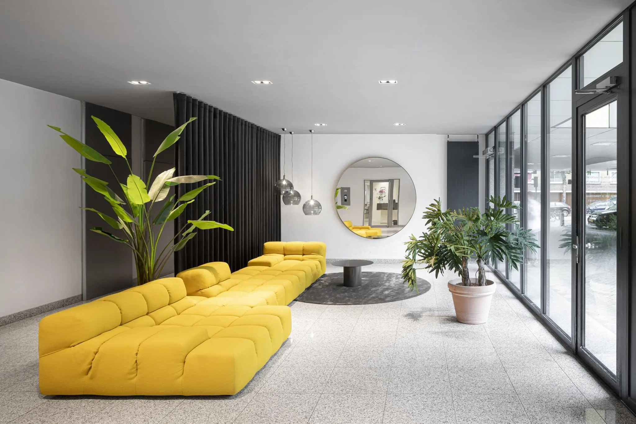 Moderne lobby aan de Van Boshuizenstraat met een gele loungebank, grote planten en een ronde spiegel aan de muur.